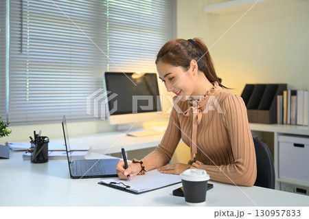 Smiling businesswoman taking notes during remote work in home workspace Smiling businesswoman taking notes during remote work in home workspace 130957833
