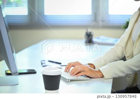 Close up of hands typing on wireless keyboard at a modern office desk with coffee cup 130957848