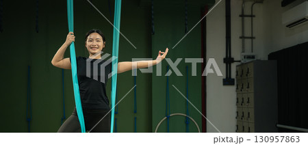 Young woman practicing aerial yoga with hammock fabric, smiling confidently while balancing Young woman practicing aerial yoga with hammock fabric, smiling confidently while balancing 130957863