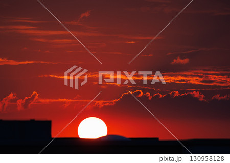 Stunning fiery sunset scene with vibrant red sky glowing sun setting behind city buildings urban silhouettes, creating warm dramatic atmosphere. Tranquil horizon dusk cloudscape Stunning fiery sunset scene with vibrant red sky glowing sun setting behind city buildings urban silhouettes, creating warm dramatic atmosphere. Tranquil horizon dusk cloudscape 130958128