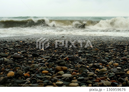 Stormy sea waves crushing on pebble beach with white foam in overcast weather. 130959676