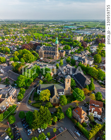 A European town with a tall green-spired Gothic church at its center, surrounded by houses, trees, and streets, glowing warmly in golden hour light. 130959755