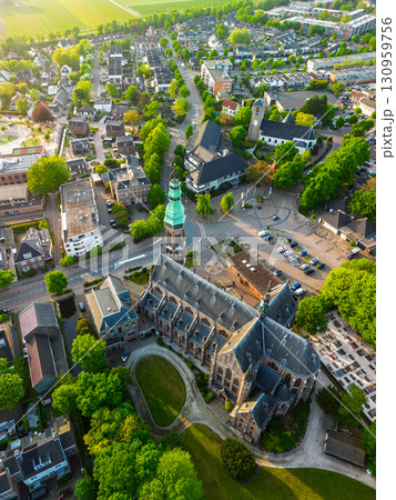 A European town with a tall green-spired Gothic church at its center, surrounded by houses, trees, and streets, glowing warmly in golden hour light. 130959756