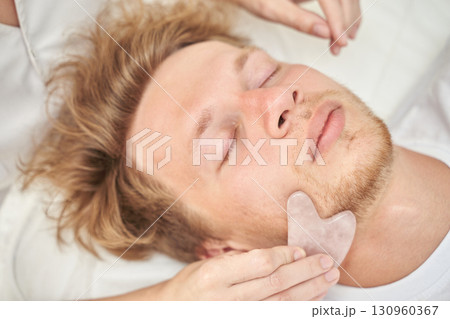 Young caucasian male relaxing with facial gua sha massage for skin rejuvenation 130960367
