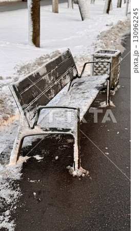 A peaceful and serene winter scene shows a snow-covered bench located in winter next to a trash can on a paved path. 130962245