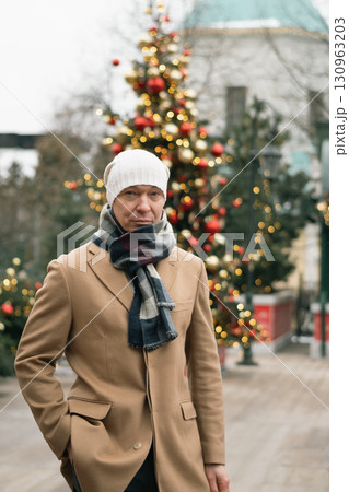 Man in cozy winter outfit stands outdoors near decorated Christmas tree, showcasing holiday decorations. Festive spirit of Xmas holiday with light garland. 130963203