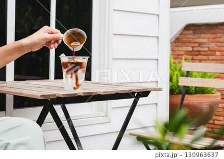 Woman pouring espreso coffee into glass with water on wooden table. Woman pouring espreso coffee into glass with water on wooden table. 130963637