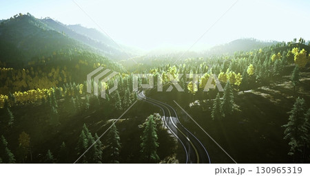 A train track winds through vibrant green forests and majestic mountains. Sunlight filters through the trees, creating a serene and picturesque landscape at sunset. 130965319