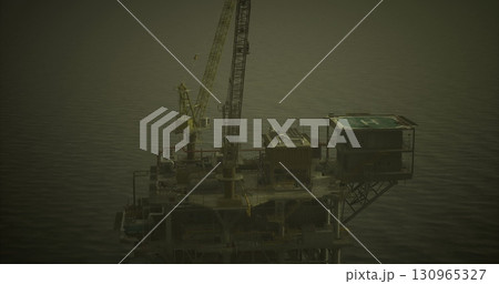 An offshore drilling platform stands tall above the water, illuminated by artificial lights as the sun sets in the distance. The calm ocean reflects the structures silhouette. An offshore drilling platform stands tall above the water, illuminated by artificial lights as the sun sets in the distance. The calm ocean reflects the structures silhouette. 130965327