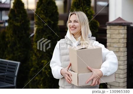 Smiling delivery woman carrying cardboard boxes in front of a house 130965380