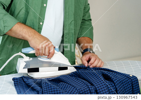 Man ironing a blue checkered shirt at home in a well-lit room Man ironing a blue checkered shirt at home in a well-lit room 130967543