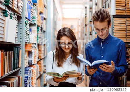 Two friends students reading books standing in the long corridor in library Two friends students reading books standing in the long corridor in library 130968325