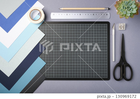 Flat lay of craft supplies including colored paper, cutting mat, scissors, ruler, pencil, masking tape, utility knife, sharpener on a gray background. Copyspace Flat lay of craft supplies including colored paper, cutting mat, scissors, ruler, pencil, masking tape, utility knife, sharpener on a gray background. Copyspace 130969172
