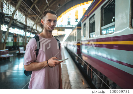 traveler guy boarding train and messaging with phone at busy railroad station, young man with backpack and smartphone starting journey on railway platform traveler guy boarding train and messaging with phone at busy railroad station, young man with backpack and smartphone starting journey on railway platform 130969781