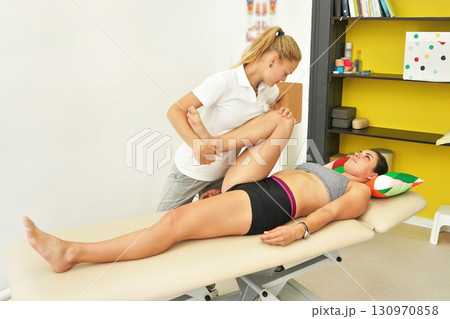 Young female physiotherapist exercising with her patient laying on massage table, flexing her knee muscles 130970858
