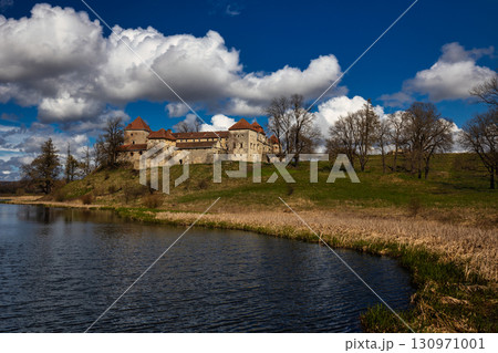 A historic stone castle with red-roofed towers stands on a grassy hill, framed by leafless trees and a calm water body, beneath dramatic clouds in a deep blue sky. 130971001