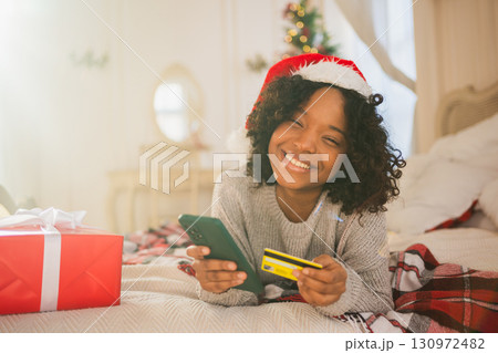 African American woman shopping online holding smartphone paying with gold credit card for Christmas gifts. Girl buying on Internet enter credit card details enjoying Christmas winter holidays at home 130972482