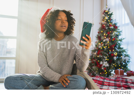 Merry Christmas. African woman in Santa hat having video chat in smartphone. Blogger in festive mood recording video in social network. Girl having video call with friends family celebrating Christmas 130972492
