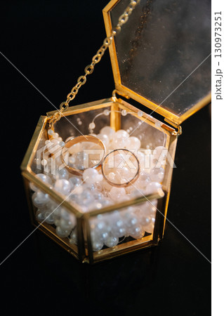 Two gold wedding rings in a glass box filled with decorative pearls on black background 130973251