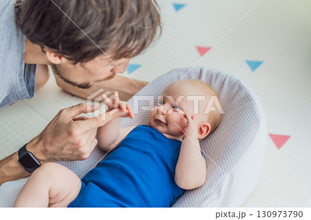 Loving father interacts with his 3-month-old baby lying in a cozy baby nest at home. Tender bonding moment, fatherhood, and early connection in a peaceful family setting Loving father interacts with his 3-month-old baby lying in a cozy baby nest at home. Tender bonding moment, fatherhood, and early connection in a peaceful family setting 130973790
