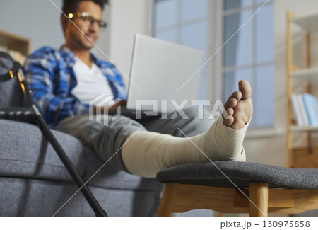 Young man with broken leg sitting on sofa using laptop Young man with broken leg sitting on sofa using laptop 130975858