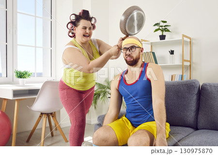 Funny, crazy housewife having fun and hitting her husband on head with frying pan Funny, crazy housewife having fun and hitting her husband on head with frying pan 130975870