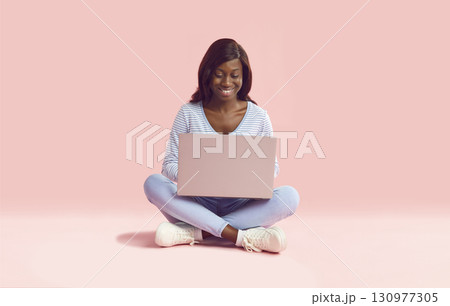 Happy young African American woman sitting on pink background and using laptop computer Happy young African American woman sitting on pink background and using laptop computer 130977305