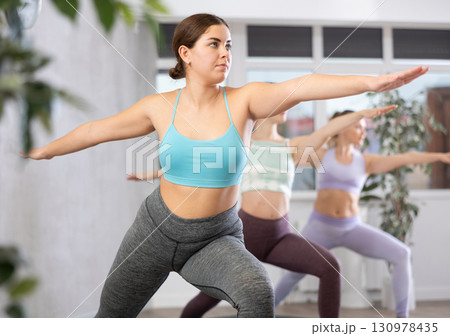 Motivated fit sports women performing Virabhadrasana 2 or warrior 2 pose on mats during yoga workout in fitness center Motivated fit sports women performing Virabhadrasana 2 or warrior 2 pose on mats during yoga workout in fitness center 130978435