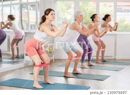 Elderly woman stands on mat and performs stretching exercises with resistance bands in group pilates class Elderly woman stands on mat and performs stretching exercises with resistance bands in group pilates class 130978897