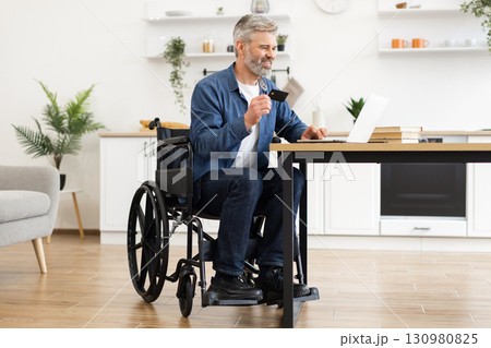 Mature man in wheelchair using laptop for online payment with bank card inside modern home setting. Concept of accessibility, financial independence, and modern technology for everyday transactions. 130980825