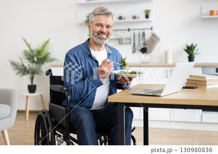 Older adult man in wheelchair eating fresh salad, working on laptop, indoors setting. Demonstrates active lifestyle, independence, healthy eating habits, home office work environment. 130980836