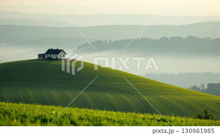 Green hill with small house on top surrounded by misty layered forest hills in background creating peaceful rural landscape 130981985
