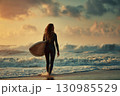 Female surfer in black wetsuit with surfboard entering sea at sunset 130985529