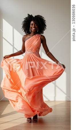 A happy woman with an afro hairstyle smiling in a beautiful peach one-shoulder evening gown 130986036