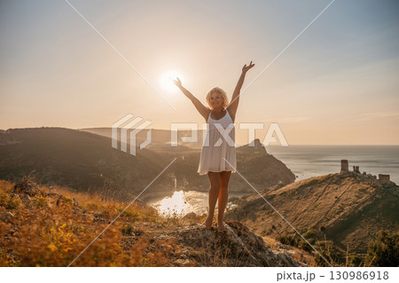 woman standing hill with her arms raised in the air, looking up at the sun. The scene is peaceful and serene, with the woman's expression conveying a sense of joy and happiness. 130986918