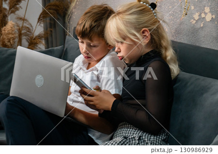 Children Laptop Technology - Two kids sit on a couch using a laptop and tablet in a home. 130986929