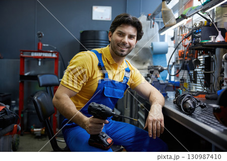 A man worker sits comfortably in a chair, holding a drill and smiling 130987410