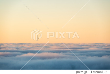 View above the clouds with the setting sun glowing on the horizon and sky painted in warm orange tones, creating a dreamy and surreal aerial sunset scene. 130988122