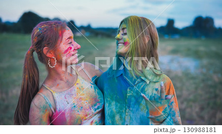 Two vibrant young women, covered in holi powder, share a joyful embrace at a colorful festival celebration Two vibrant young women, covered in holi powder, share a joyful embrace at a colorful festival celebration 130988181