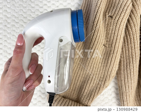 A woman holds a lint remover against a warm sweater with bobbles, removing pilling and caring for clothes. Preparing wardrobe for winter and autumn seasons, clothing care and maintenance 130988429
