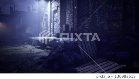 An eerie alleyway is illuminated by a lone streetlight at night. Piles of debris, including wooden pallets and refuse, create a haunting atmosphere in the dimly lit space. An eerie alleyway is illuminated by a lone streetlight at night. Piles of debris, including wooden pallets and refuse, create a haunting atmosphere in the dimly lit space. 130988615