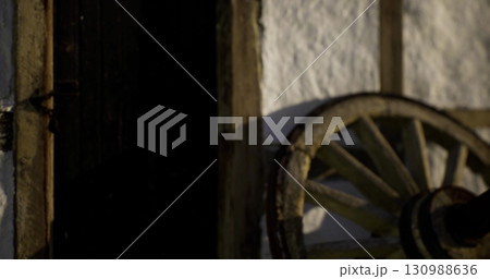 A weathered wooden wheel rests beside a rustic door, casting shadows on the textured wall. The warm light of sunset enhances the villages historic charm and tranquility. 130988636