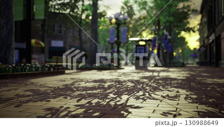 Golden sunlight filters through trees, creating intricate leaf shadows on the paved walkway in a serene urban park during evening hours, ideal for relaxation and socializing. 130988649