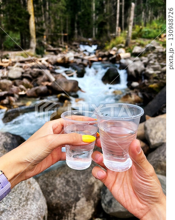 Two hands holding glasses of clear water, with a mountain river in the background. 130988726