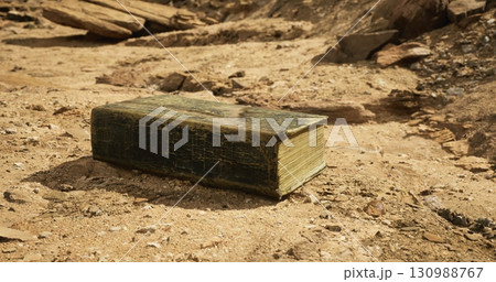 An old book lies motionless on parched earth, surrounded by rocky terrain under a bright sky, signifying remnants of history amidst a barren environment. An old book lies motionless on parched earth, surrounded by rocky terrain under a bright sky, signifying remnants of history amidst a barren environment. 130988767