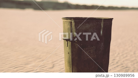 A rusted trash can, weathered and worn, stands solitary on a vast expanse of soft sand at dusk, with golden light casting long shadows. Nature slowly reclaims its space and beauty. A rusted trash can, weathered and worn, stands solitary on a vast expanse of soft sand at dusk, with golden light casting long shadows. Nature slowly reclaims its space and beauty. 130989956