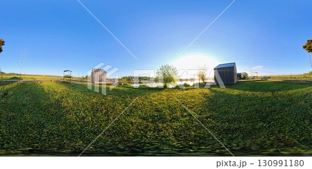 360-degree panoramic HDRI image of a serene countryside scene at sunrise. A wooden boardwalk stretches between grassy fields and small lakeside cabins. 130991180