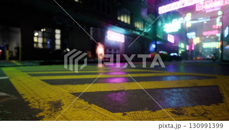 A vibrant urban street is bathed in neon lights reflecting off wet pavement at night. The atmosphere is lively with bright signs and city energy in the background. 130991399
