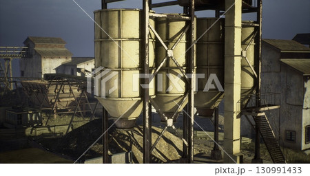 Large silos are positioned on a construction site surrounded by machinery and old buildings. The setting showcases industrial operations under clear skies during the day. 130991433