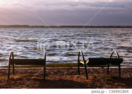Two empty benches sit on the sandy shore, facing a tranquil lake. Soft waves ripple under warm golden sunlight as clouds drift lazily above 130991804